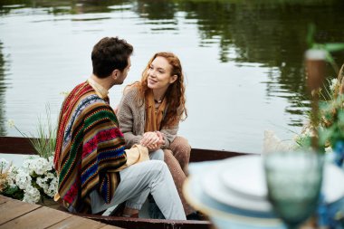 A man and woman in boho attire sit peacefully in a boat on a tranquil lake in a lush green park setting. clipart