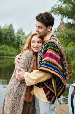 A man and woman in boho attire stand beside a tranquil body of water in a beautiful park setting. clipart