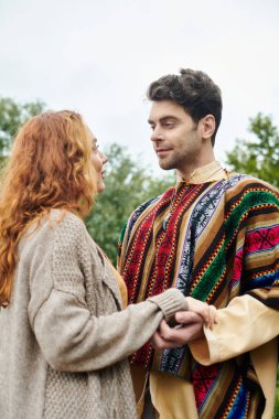 Boho tarzı giyinmiş bir erkek ve kadın, yemyeşil bir parkta yan yana durur, sevgi ve bağ hissi yayar..