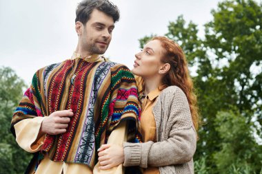 Boho tarzı giyinmiş bir erkek ve bir kadın, yeşil bir parkta yan yana durur, sevgi ve beraberlik yayarlar..