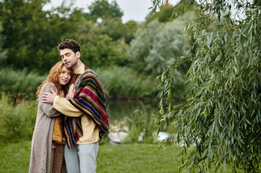 Boho tarzı giyinmiş bir kadın ve bir erkek, yeşil bir parkta birbirine yakın durur. Romantizm ve zarafet dolu bir hava yayarlar..