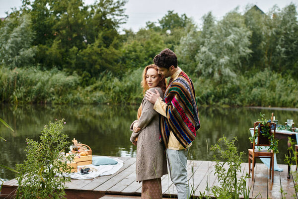 A man and a woman, dressed in boho style clothes, embrace lovingly on a dock in a green park on a romantic date.