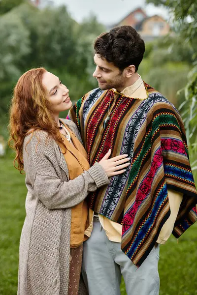 stock image A man in boho style clothing stands beside a woman wrapped in a blanket in a serene green park setting.