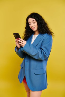 Mavi ceketli genç bir kadın cep telefonuyla büyülenmiş, yoğun bir şekilde ekrana odaklanmış..