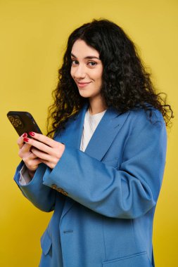 Mavi ceketli genç bir kadın cep telefonu ekranına odaklanmış, sıradan bir ortamda dalgın dalgın bir ifadeyle ayakta duruyor..