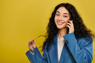 Şık mavi ceketli, sarı arka planda cep telefonuyla konuşan esmer bir kadın..