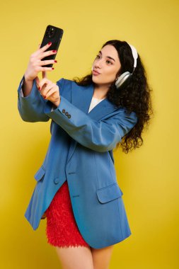 Brunette woman with curly hair in stylish attire poses for a selfie with cell phone, showing various emotions against a yellow studio backdrop. clipart