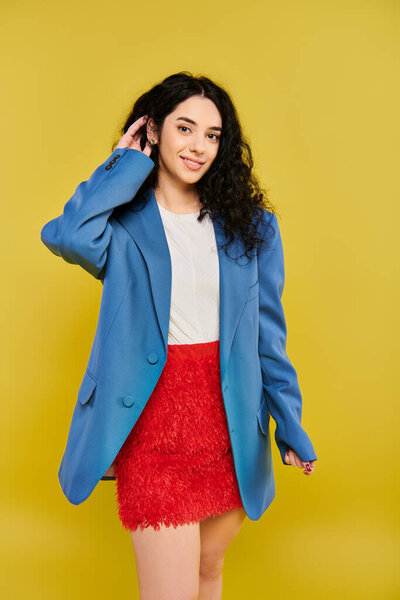 Young brunette woman with curly hair poses in stylish blue jacket and red skirt, exuding confidence and style on yellow background.