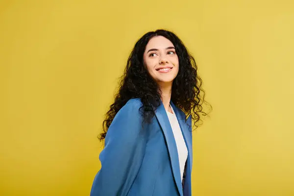 stock image A young, brunette woman in a blue jacket stands confidently in front of a striking yellow wall, exuding style and emotion.