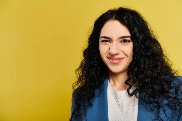 stock image A young woman with long, curly black hair wearing a stylish blue blazer, exuding confidence and elegance in a studio set with a yellow background.