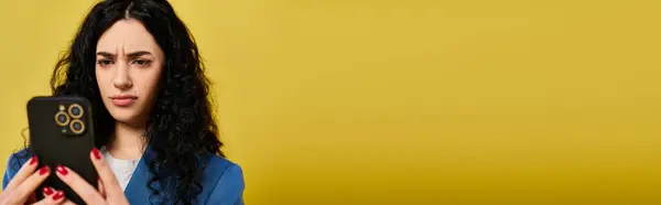 stock image Brunette woman with long, curly hair looking intently at her cell phone, showcasing a mix of emotions, against a vibrant yellow backdrop.