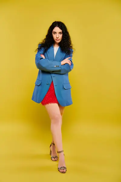 stock image A young, curly-haired brunette woman exudes confidence in a blue jacket and red skirt against a vibrant yellow background.