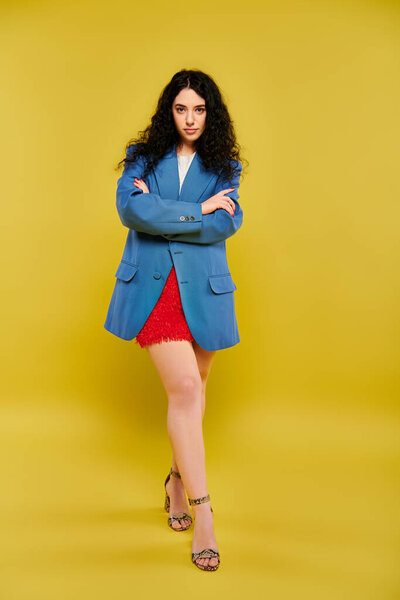 A young, curly-haired brunette woman exudes confidence in a blue jacket and red skirt against a vibrant yellow background.