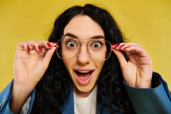 stock image A young, stylish woman with curly hair wearing glasses is expressing surprise against a vibrant yellow backdrop.