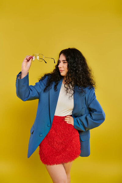 A stylish young woman with curly brown hair strikes a pose in a vibrant blue jacket and a striking red skirt against a sunny yellow backdrop.