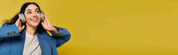 stock image A young brunette woman with curly hair wearing a blue jacket, listening to headphones against a yellow background.