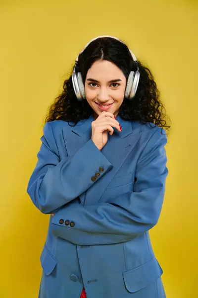 黄色い背景に対してヘッドフォンとスタイリッシュな青いスーツを着ている間 カーリーな髪の毛を持つ若いブルネットの女性 — ストック写真