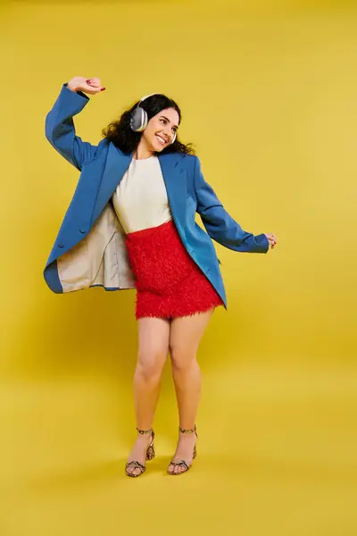 明るい黄色の背景に対してスタイリッシュな青いジャケットと赤いスカートでカーリーヘアを発射する若いブルネットの女性 — ストック写真