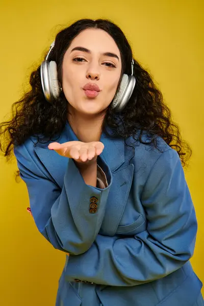 stock image A young, brunette woman with curly hair makes a funny expression while wearing headphones in a studio with a yellow background.