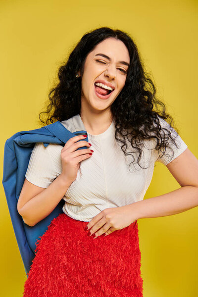 Young, curly-haired brunette woman poses with emotion in a stylish red skirt and white shirt against a vibrant yellow background.