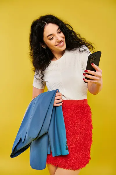 stock image Brunette woman with curly hair in red skirt poses with cell phone in hand, emoting in a studio setting with yellow backdrop.