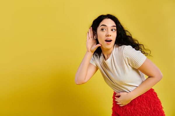 A brunette woman with curly hair looking surprised, dressed in stylish attire, in a studio with a yellow background.