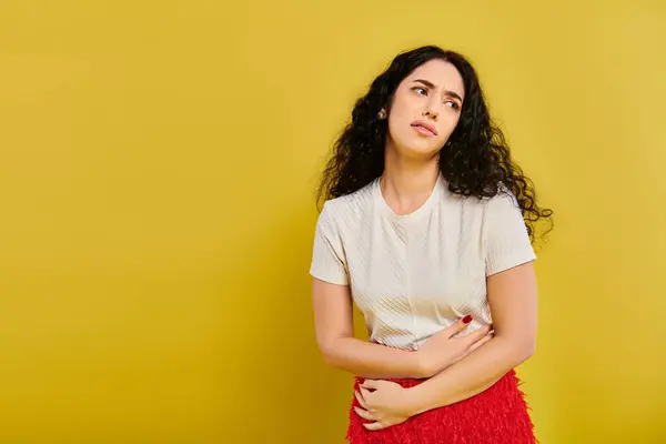 stock image Stylish young woman with curly hair posing in white shirt and red skirt in front of yellow wall, showcasing her bored face expression