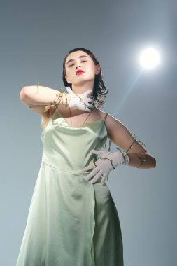 A beautiful young woman with red lips poses in a green dress and white gloves in a studio setting against a grey backdrop. clipart