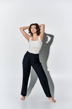 Tarz sahibi genç bir kadın, beyaz atlet bluzu ve siyah pantolonuyla bir stüdyo ortamında gri bir arkaplan karşısında poz veriyor..