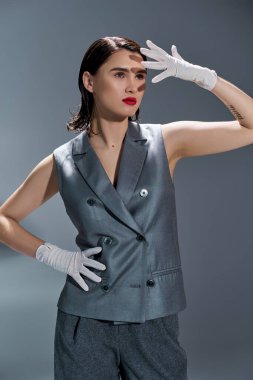 Stylish young woman striking a pose in an elegant gray suit with a vest, complemented by white gloves, on a gray studio background. clipart
