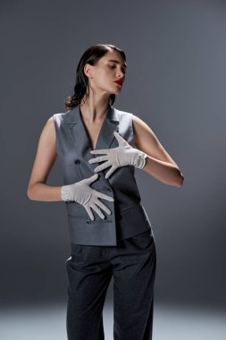 Stylish young woman strikes a pose in an elegant gray suit with a vest, complemented by white gloves, in a studio against a grey backdrop. clipart