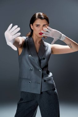 A stylish young woman is striking a pose in an elegant gray suit with a vest, complemented by white gloves, in a studio with a grey background. clipart