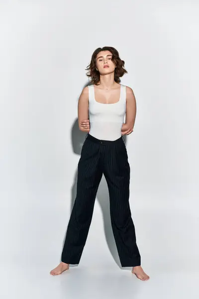 Stock image A pretty young woman in black pants and a white tank top stands confidently against a clean white wall in a studio setting.