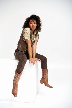 Zarif giyinmiş, beyaz küpün üzerinde oturan ve kameraya bakan Afro-Amerikan kadını cezbediyor.