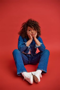 beautiful young african american woman in stylish denim outfit looking away on red backdrop clipart