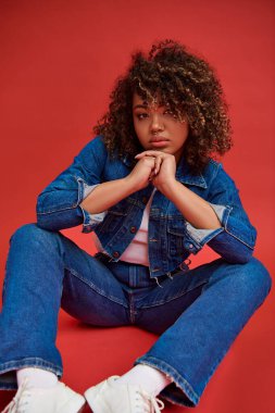 Kot pantolon giymiş süslü Afro-Amerikan kadın model kameraya bakıyor ve kırmızı arka planda poz veriyor.