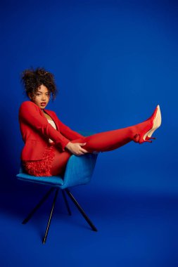 Baştan çıkarıcı afro-amerikan kadını. Kırmızı takım elbiseli, mavi koltukta oturmuş kameraya bakıyor.