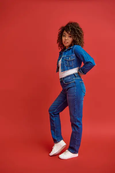 stock image fashionable african american woman in denim attire posing on red backdrop and looking at camera