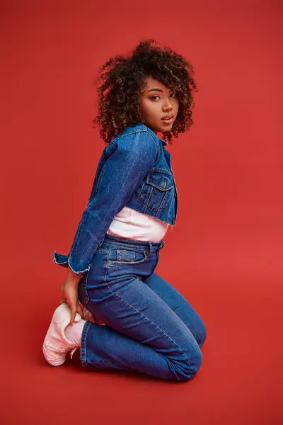 stock image tasteful african american woman in stylish denim outfit looking at camera on vibrant red backdrop