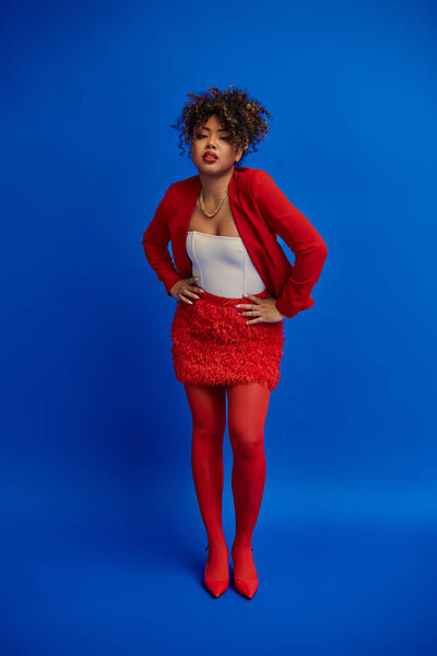 stylish african american woman in red skirt and blazer posing on blue backdrop and looking at camera