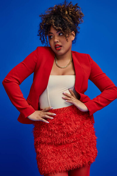 attractive stylish african american fashionista in elegant outfit looking away on blue backdrop
