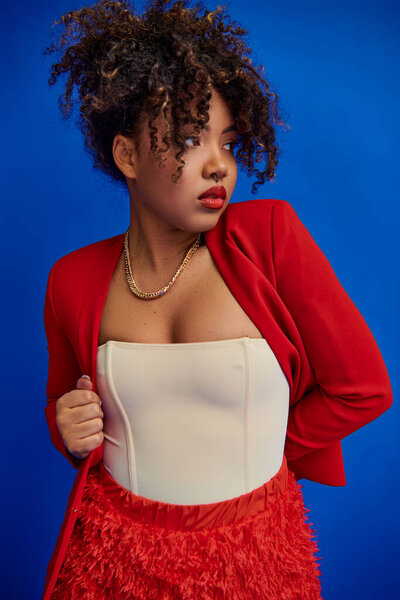 beautiful stylish african american fashionista in vivid elegant outfit looking away on blue backdrop