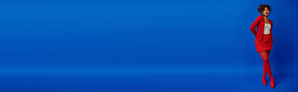 young african american woman in red blazer with curly hair looking at camera, blue backdrop, banner