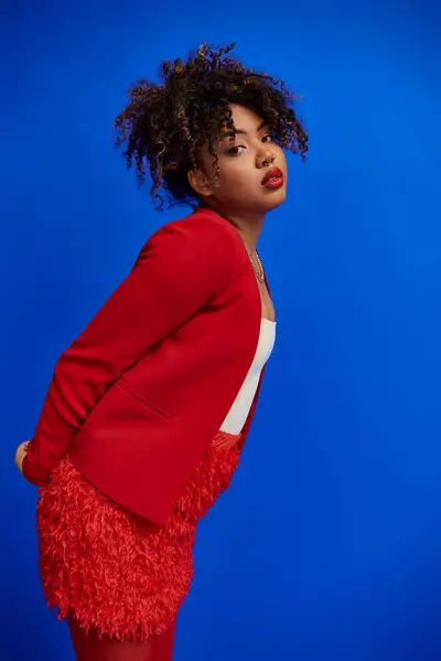 カメラ 青い背景を見ているカーリー髪の赤いブレーザーの優雅なシックなアフリカ系アメリカ人女性 — ストック写真