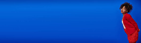 chic african american woman in red blazer with curly hair looking at camera, blue backdrop, banner