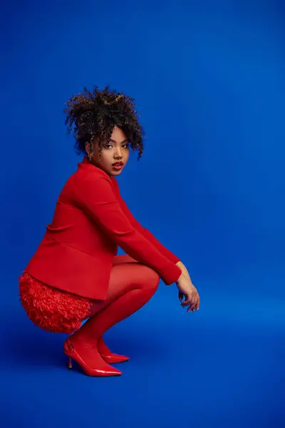 stock image fancy chic african american woman in red blazer with curly hair looking at camera, blue backdrop