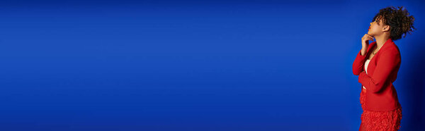 chic african american woman in vivid attire with collected hair looking away, blue backdrop, banner