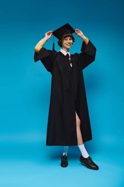 Stüdyoda mavi arka planda duran siyah mezuniyet elbisesi ve şapkası giyen mutlu üniversiteli kız.