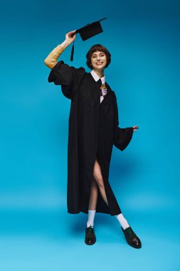Stüdyoda mavi arka planda akademik şapkası olan siyah mezuniyet elbisesi giyen mutlu üniversiteli kız.