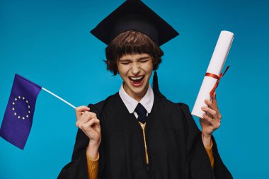 Mavi arka planda diploma ve AB bayrağı taşıyan, önlüklü ve akademik şapkalı üniversite mezunu heyecanlı kız